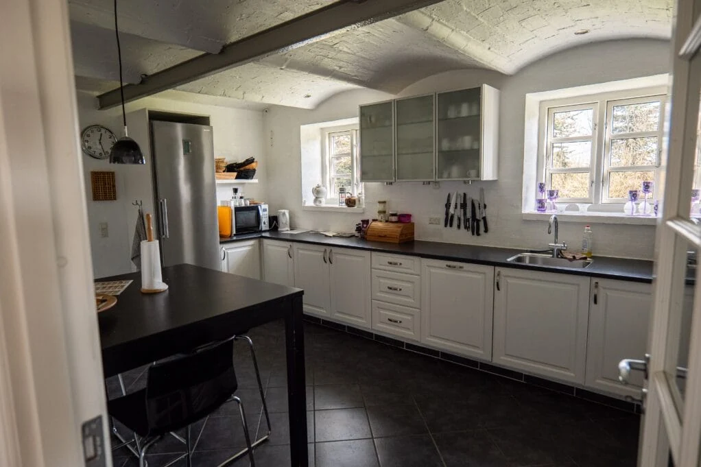 Køkkenet med hvide låger, mørk bordplade, køleskab og vask under vinduerne.